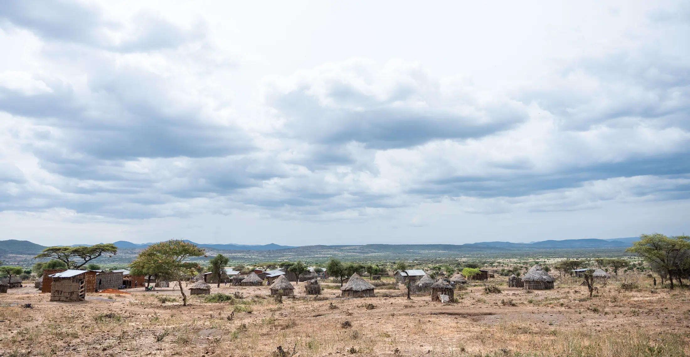 Wide view of a rural settlement under a cloudy sky, round huts scattered across dry grassland with hills beyond.