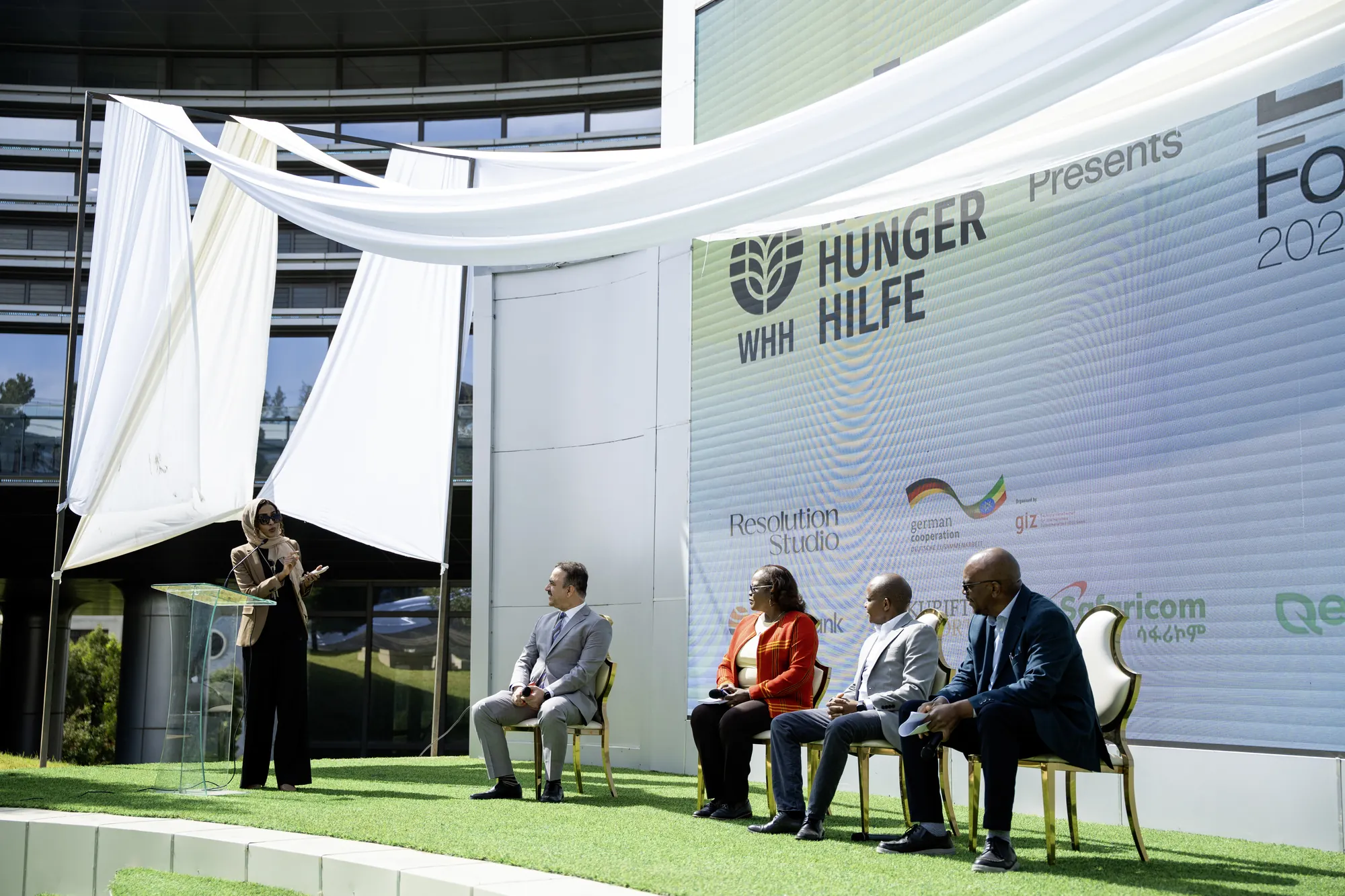 The panel on stage under the white canopy, the audience in the foreground.