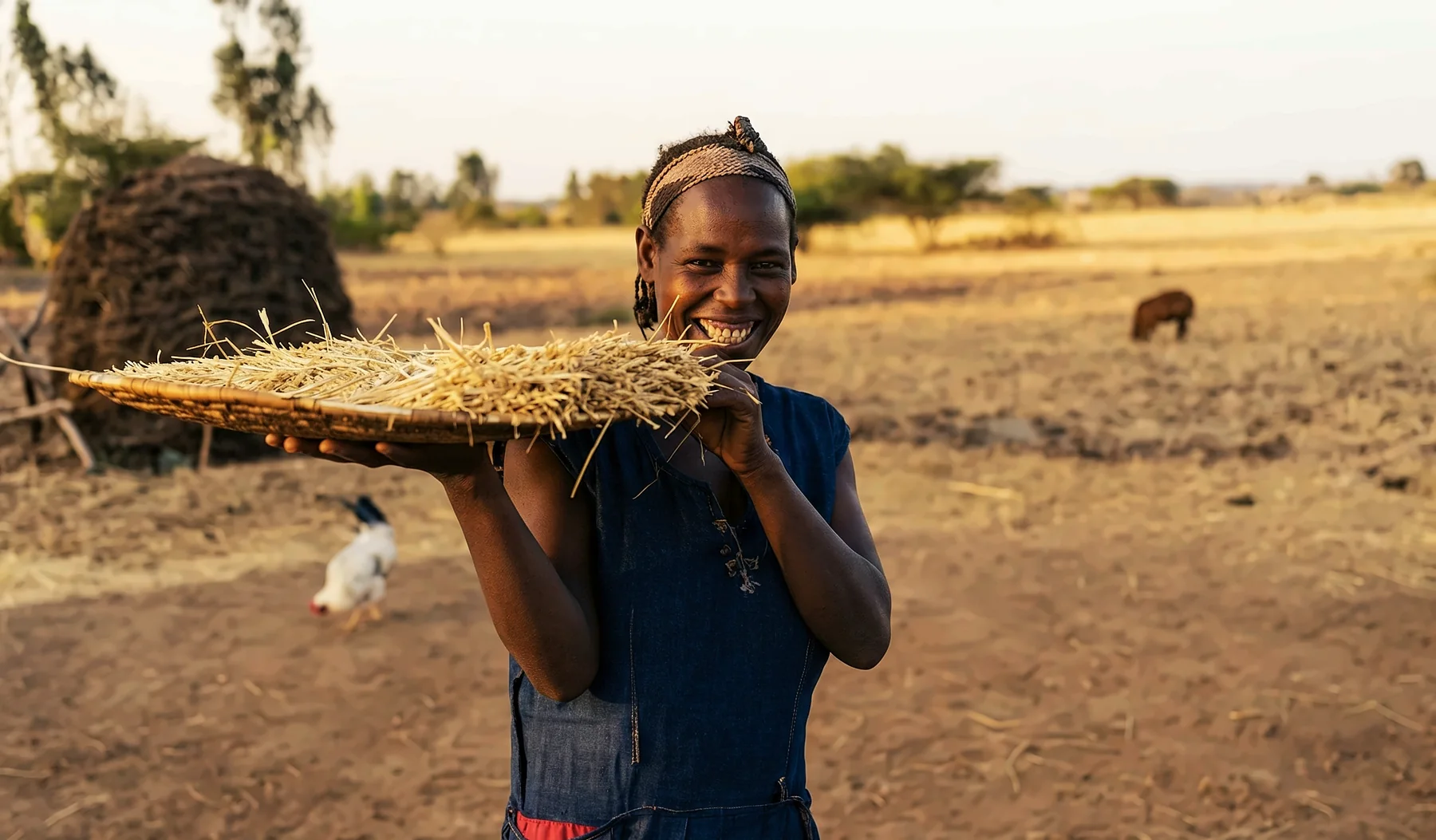 A smiling woman holding a woven tray of straw in a dry field at dusk.