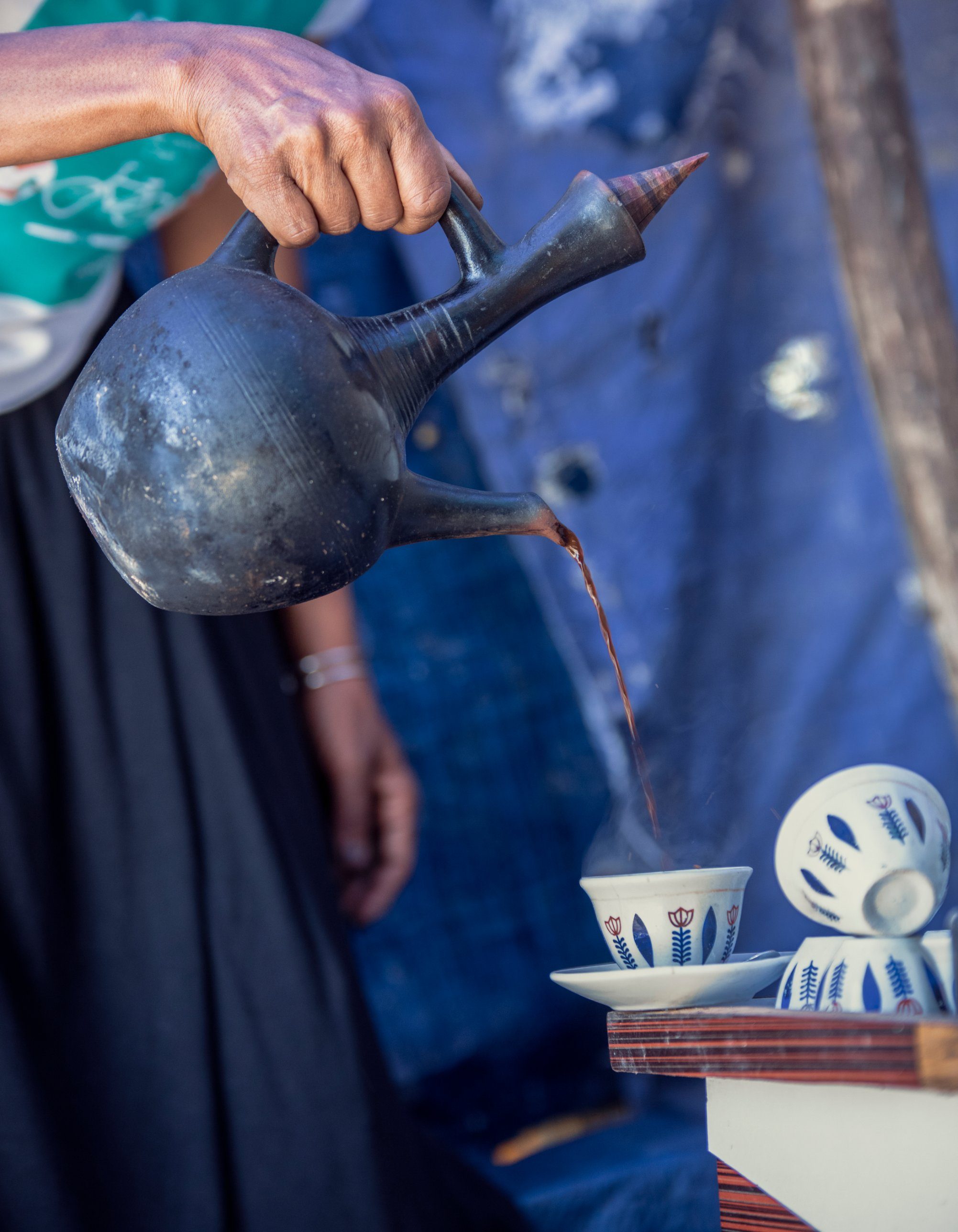A coffee vendor pours steaming coffee from a clay jebena into a small painted cup.
