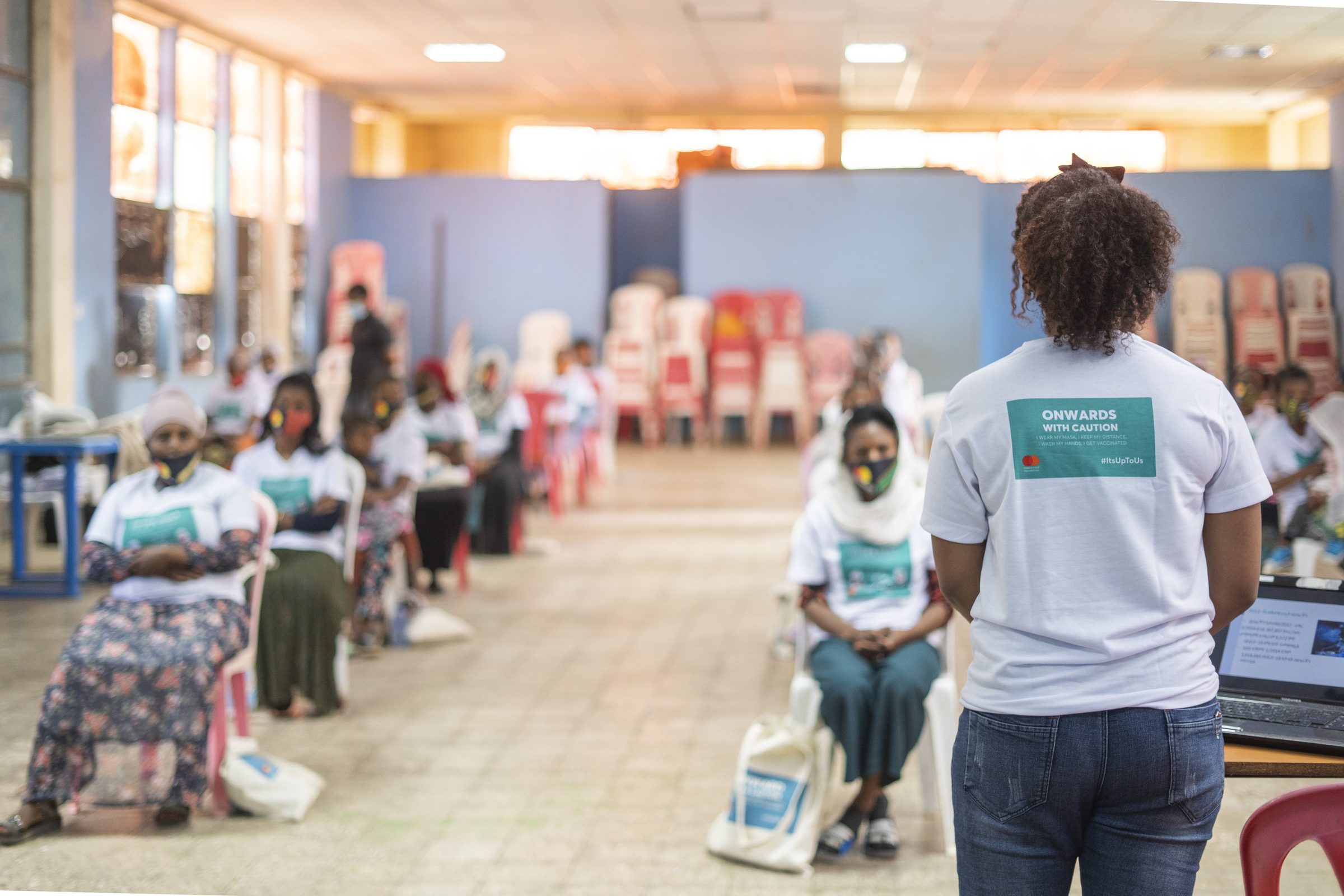 A trainer stands at the front of a hall of seated coffee vendors in campaign T-shirts.