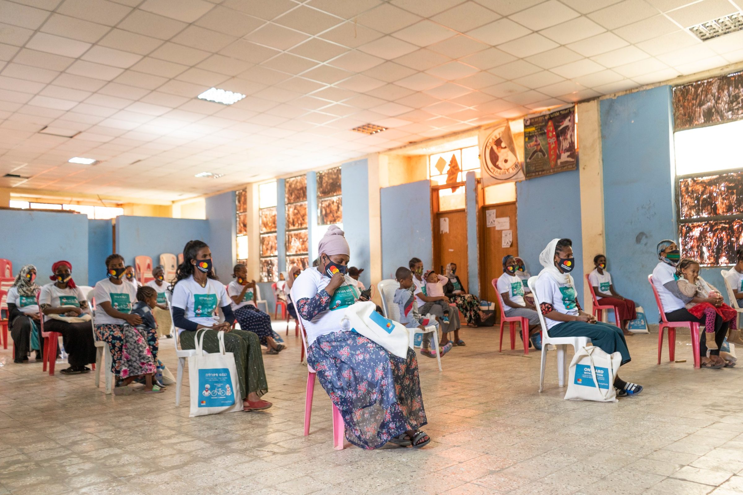 Coffee vendors seated in a hall, campaign tote bags at their feet.