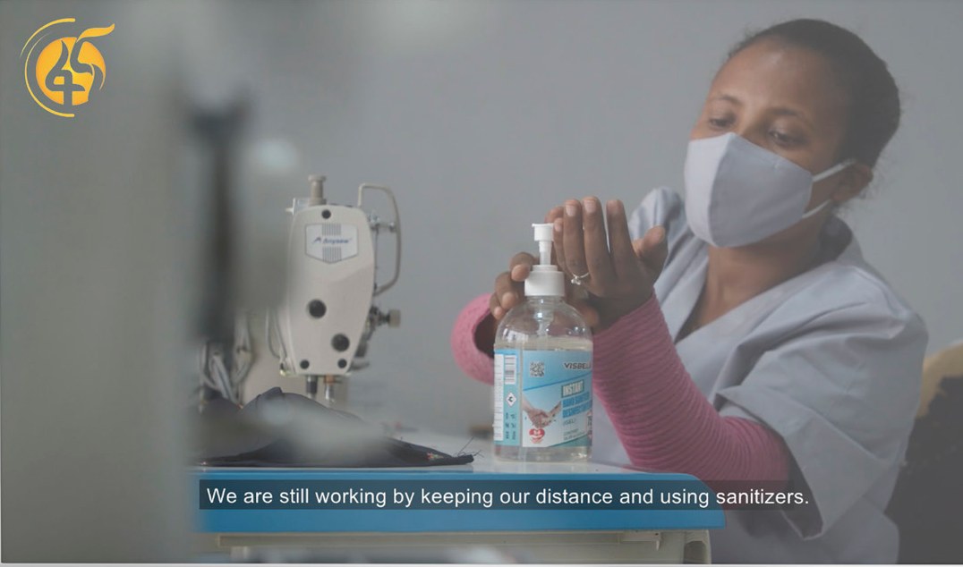 TV spot still: a masked seamstress uses hand sanitizer at her sewing machine. Subtitle: We are still working by keeping our distance and using sanitizers.