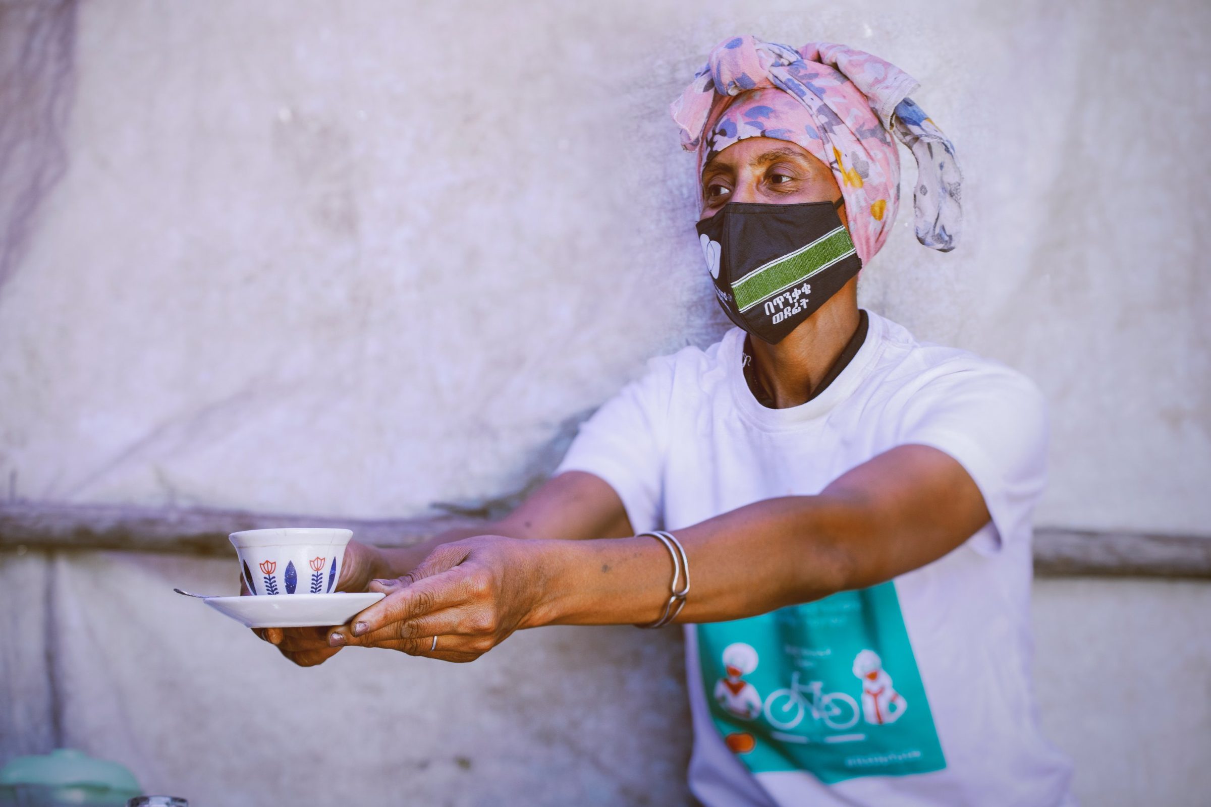 A coffee vendor in an Onward with Caution mask and campaign T-shirt holds out a cup of coffee on a saucer.