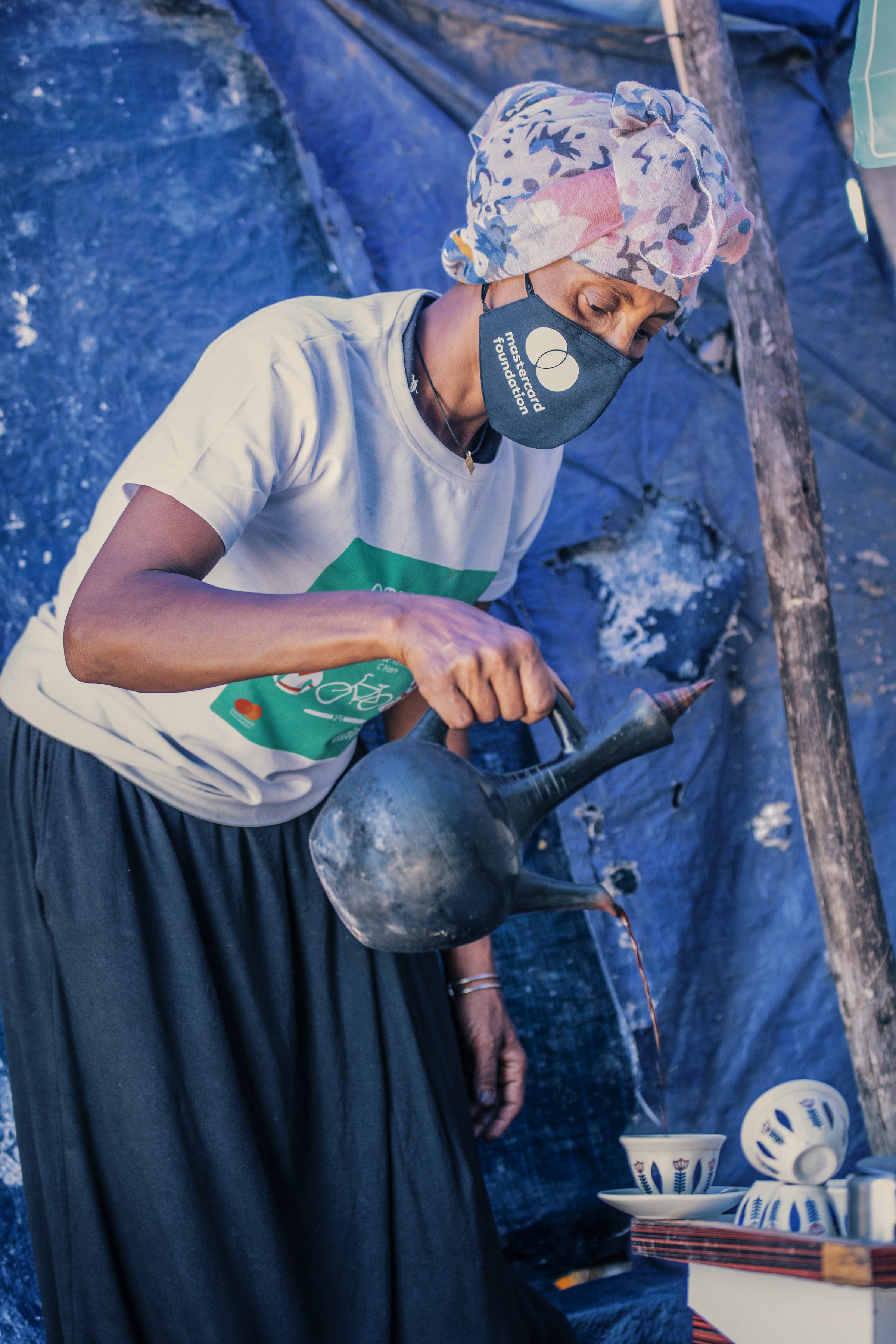 A coffee vendor in a mask pours from a jebena at her stall under blue tarpaulin.